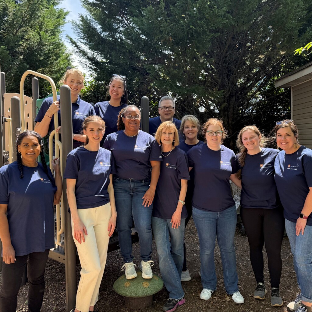 A group of Dority & Manning volunteers pose together outdoors in front of a playground structure during the firm’s first annual Day of Giving. The group, wearing matching navy blue volunteer t-shirts, stands smiling in a sunny area surrounded by trees and a building, reflecting a spirit of community service and teamwork.
