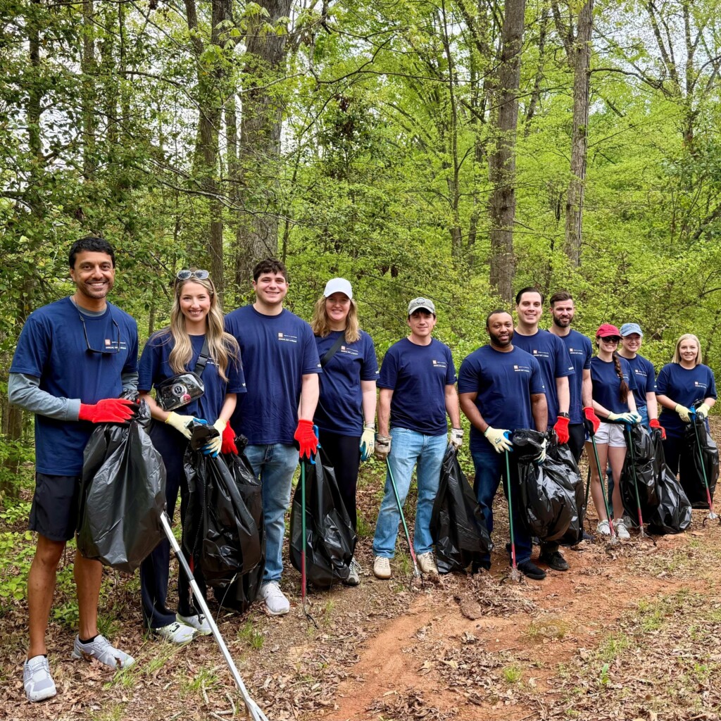 A group of Dority & Manning volunteers stand together on a forest trail, holding trash bags and litter pickers during the firm’s first annual Day of Giving. They are wearing matching navy blue volunteer t-shirts and red gloves, surrounded by lush green trees, as they participate in a community clean-up effort with smiles and teamwork.